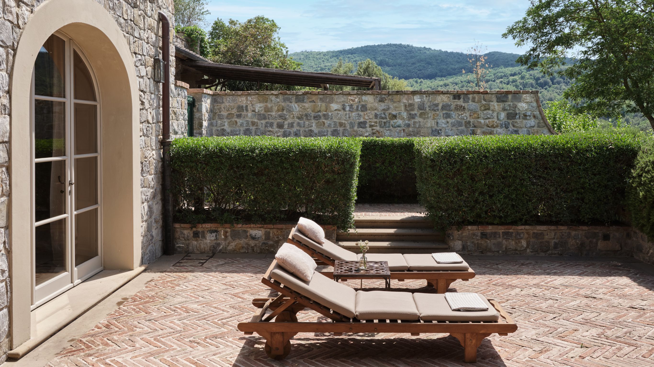 Two wooden lounge chairs with cushions sit on a brick patio under the Tuscan sun